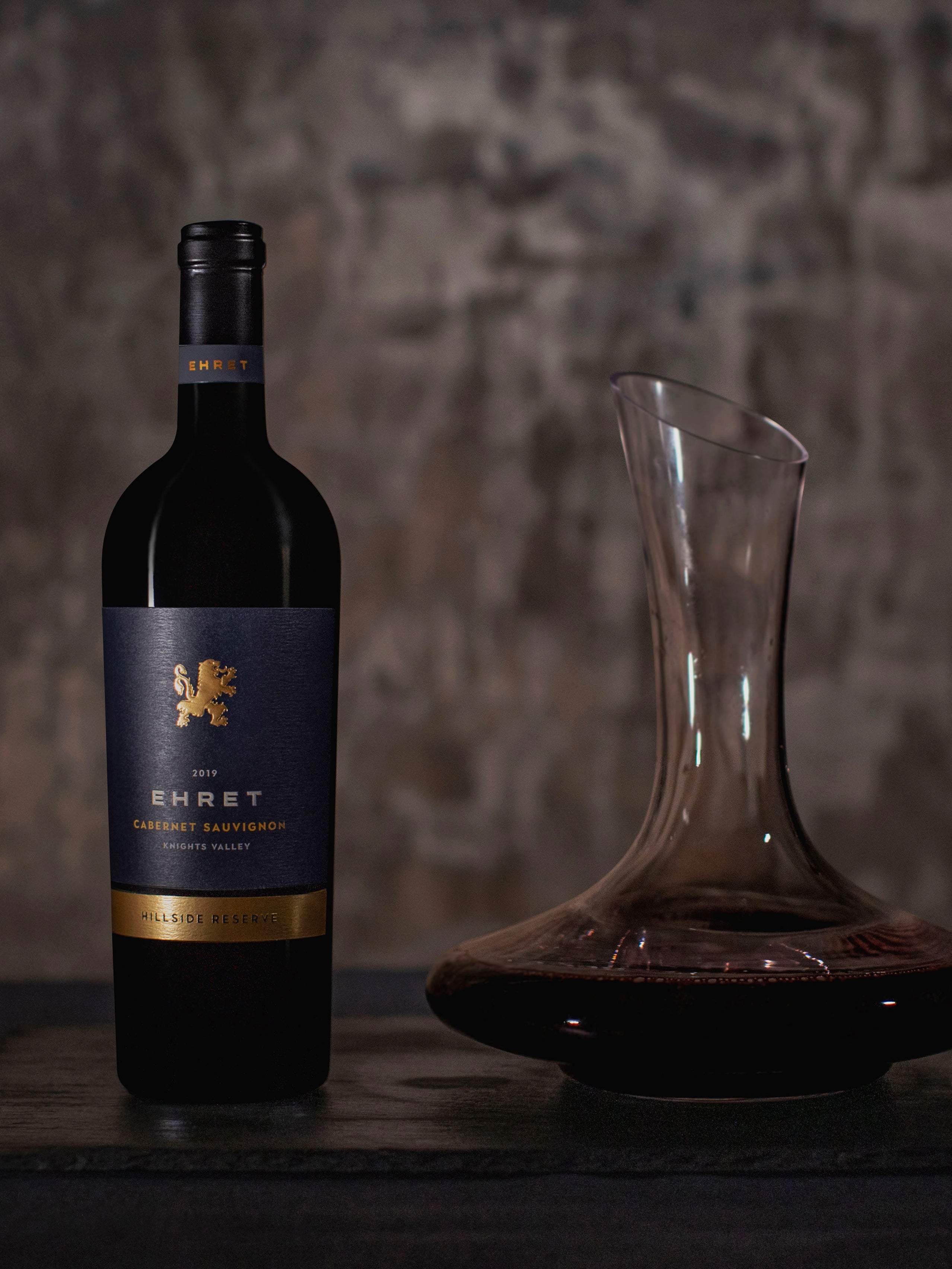 A bottle of Ehet Cabernet Sauvignon red wine stands on a dark surface next to a clear glass decanter with some red wine, set against a textured, rustic gray-brown background.