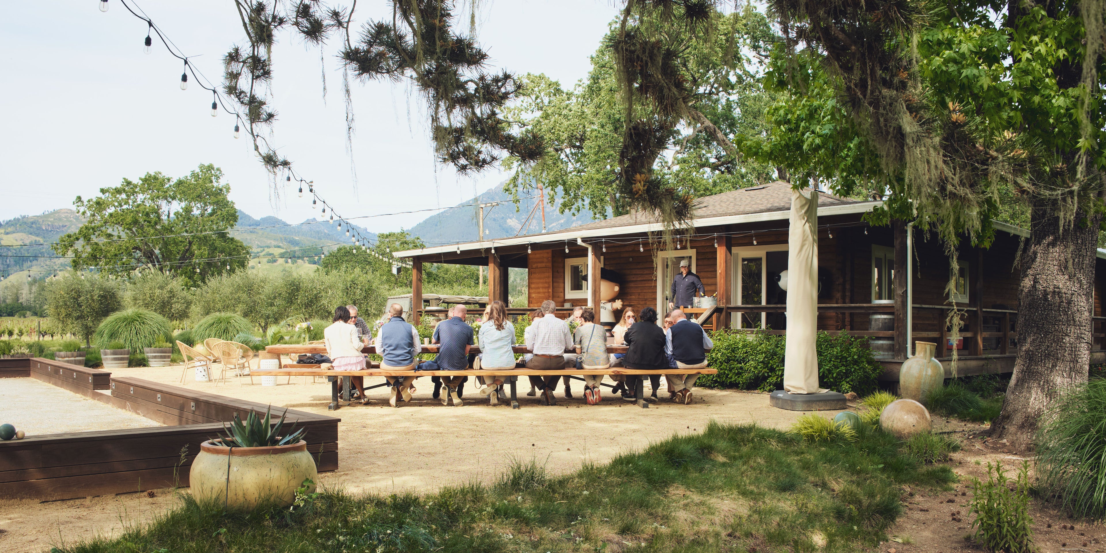 A group of people sitting on wooden tables on a patio in front of a rustic cabin celebrating a private event with Ehret Winery, surrounded by trees with hanging moss and a mountain in the background.