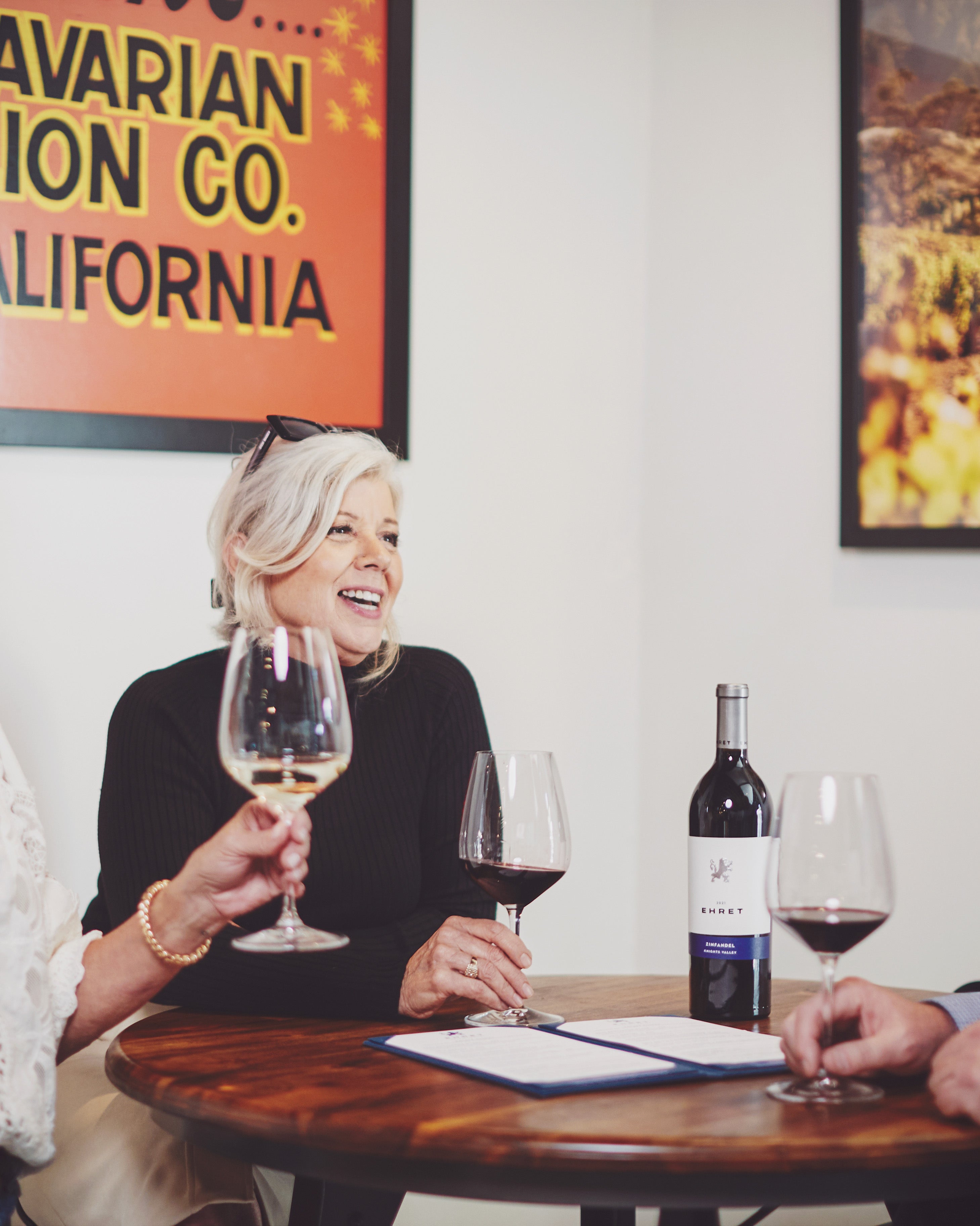 A woman with blonde hair wearing a black top smiles while holding a wine glass at a round wooden table at Ehret Winery Tasting Room Healdsburg. Other wine glasses, a bottle of Ehret Zinfandel red wine, and a menu are on the table.