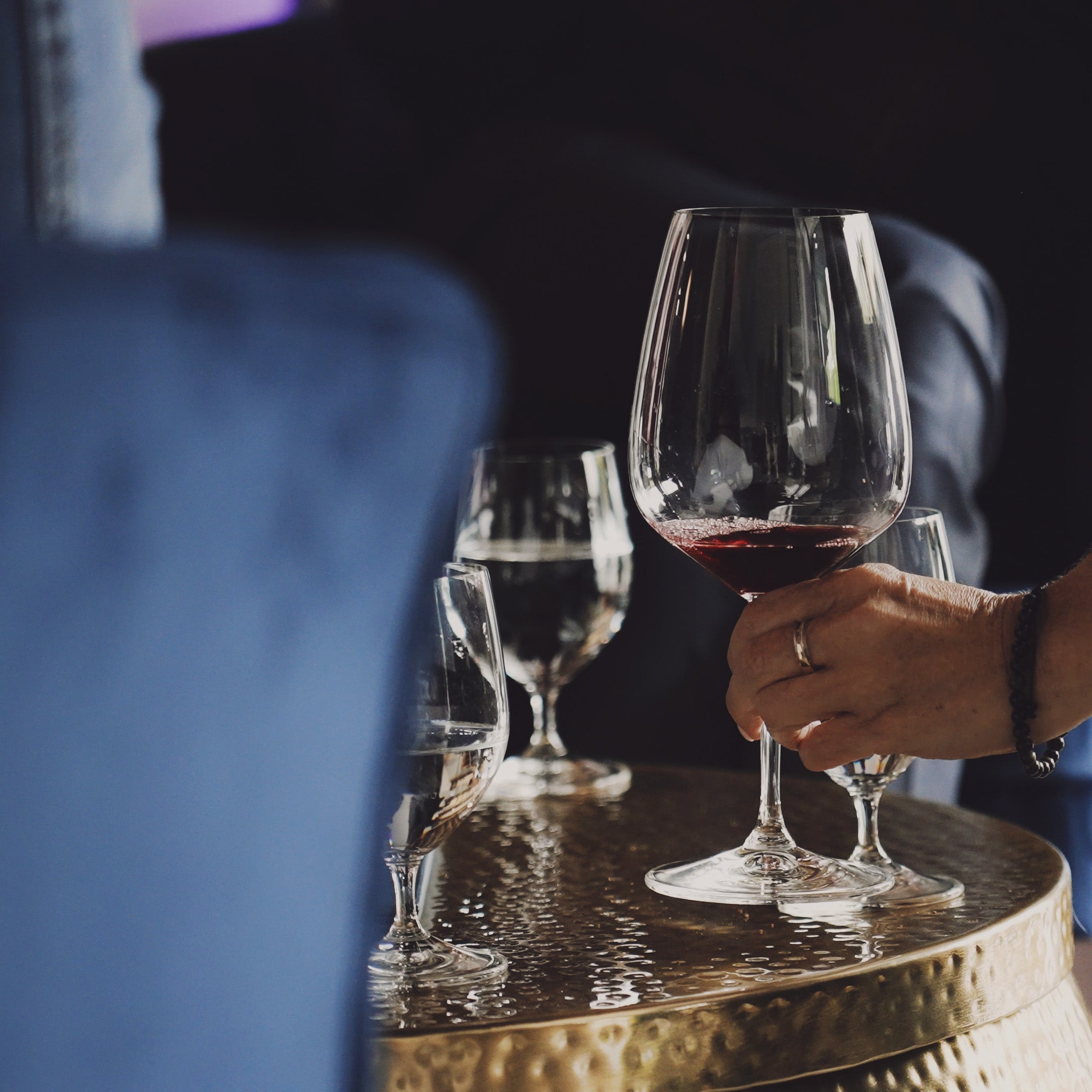 A close-up lifestyle shot of a hand holding a Ehret Winery wine glass with red wine, set on a hammered gold side table next to other glasses, with a blurred blue chair in the foreground.
