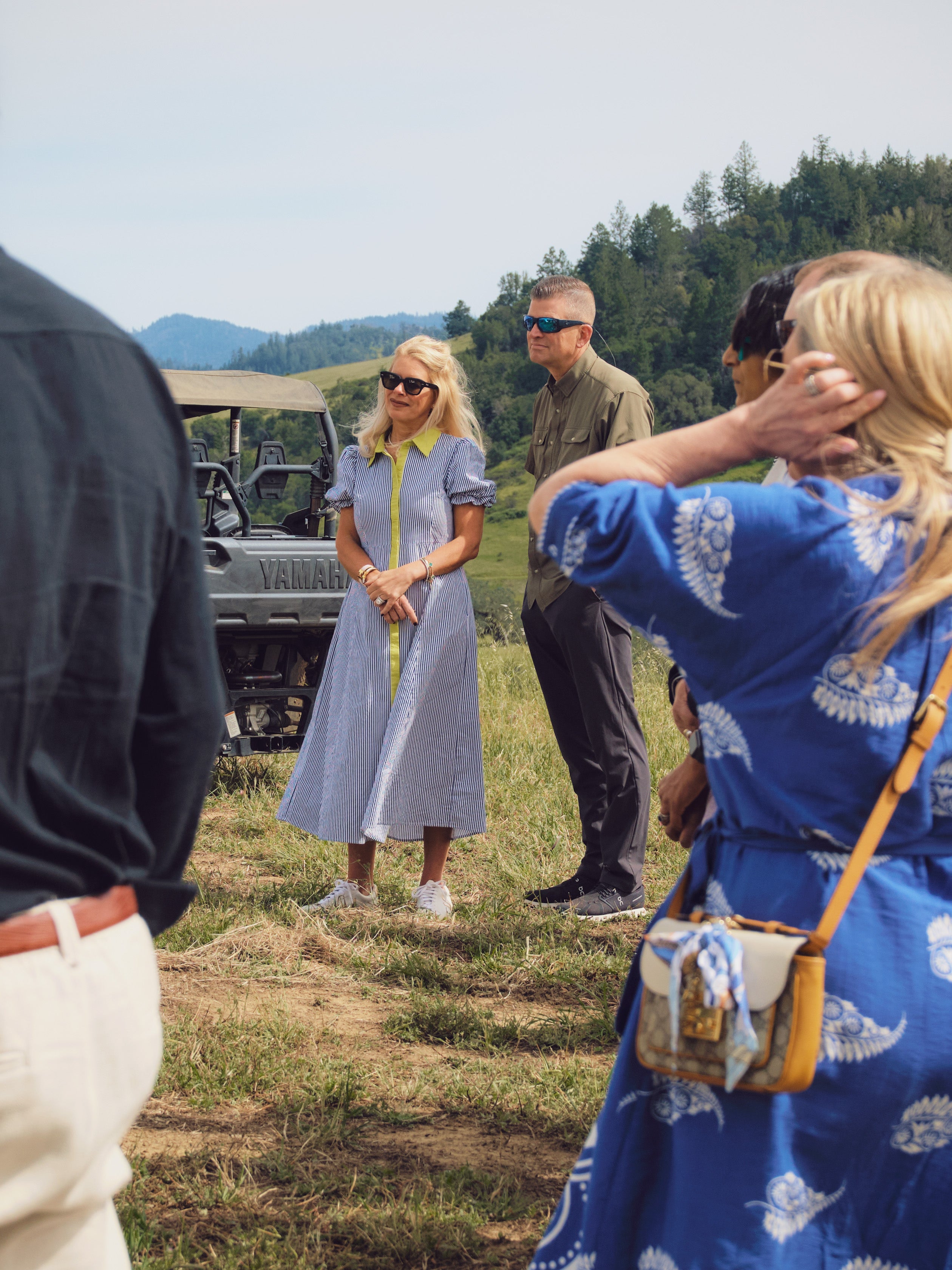A woman in a light blue and white gingham midi dress with a yellow collar stands on top of hill on Bavarian Lion Vineyards in Knights Valley Calistoga outdoors in a grassy field near a vehicle, surrounded by other people in a beautiful environment.