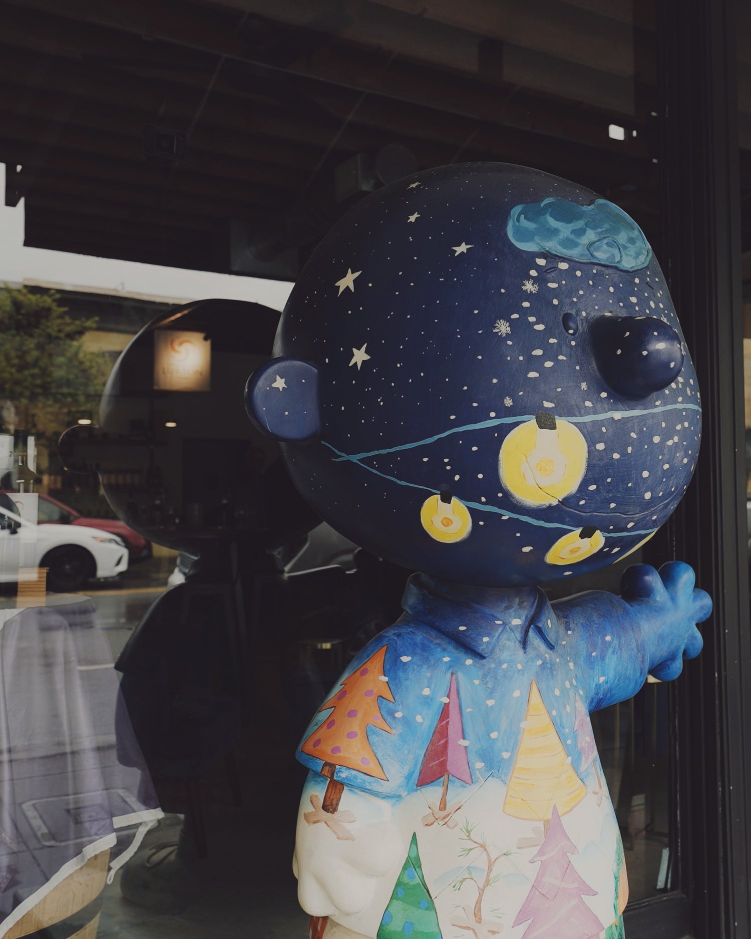 A large, stylized bear-shaped sculpture or collectible figure with a dark blue night-sky pattern on its head and a blue shirt decorated with colorful Christmas trees, photographed in front of Ehret Winery Tasting Room Healdsburg setting with reflections and a dark interior visible.