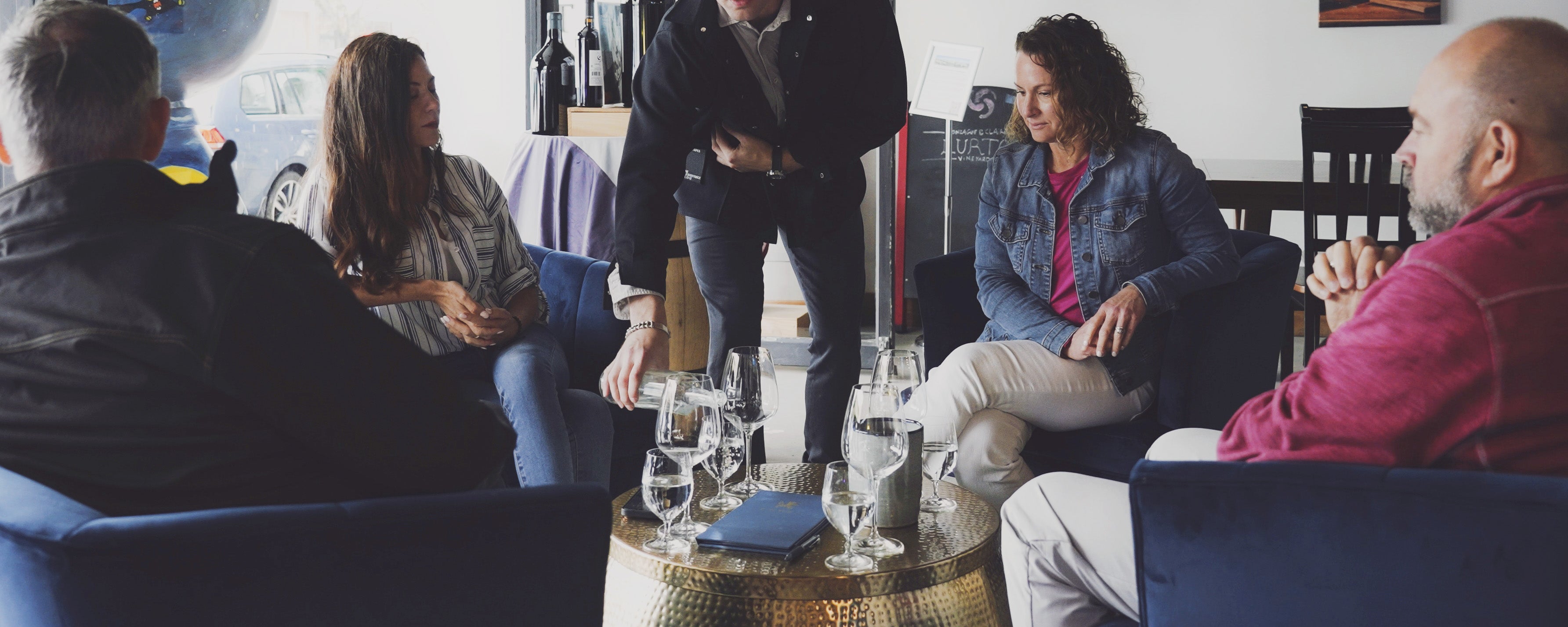 A group of five people gathered in a lounge or bar setting, seated at a round table with glasses and a bottle while Christian Ehret is pouring White Wine. The scene is indoors with blue upholstered seating and dim ambient lighting in the Ehret Winery Tasting Room Healdsburg.