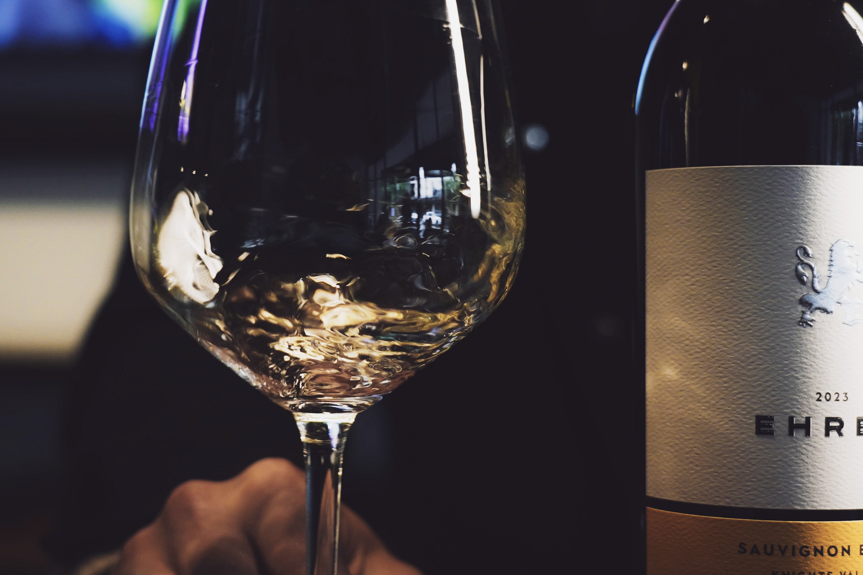 A close-up shot of a wine bottle next to a large wine glass with white wine, with a hand holding the stem. The bottle label is partially visible and reads '2023 EHRET Winey SAUVIGNON Blanc'.