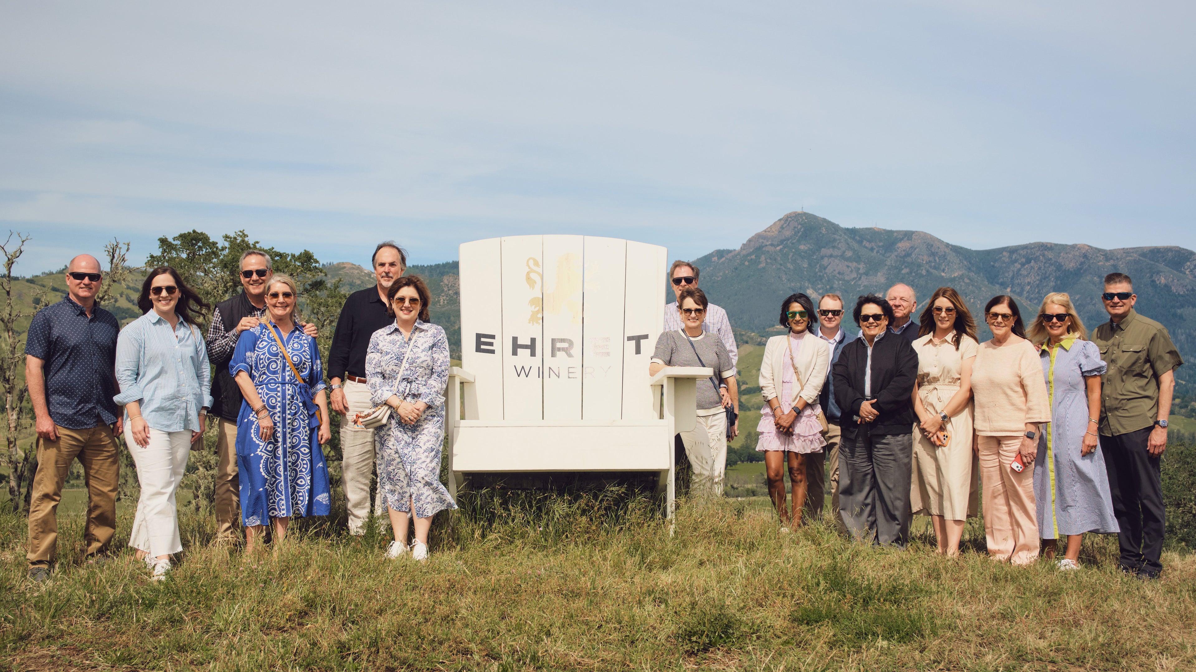 A group of people standing outdoors in a grassy hill on the Bavarian Lion Vineyards with mountains in the background, posing happy in front of a white sign that reads 'Ehret Winery'.