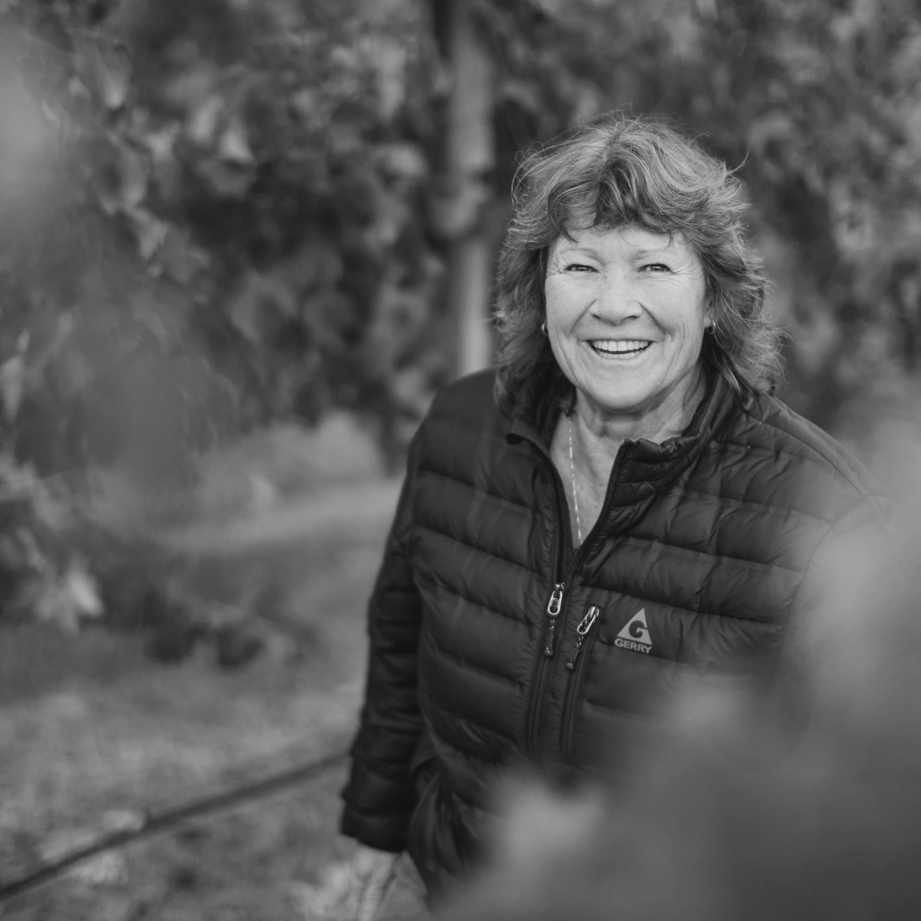 A black and white portrait of a smiling woman Erin Green Winemaker in a vineyard wearing a puffer jacket, with blurred grapes in the background.